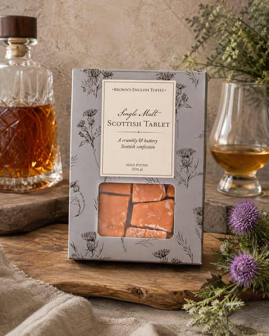 A boxed confection product labeled 'Simple Malt Scottish Tablet' stands on a rustic wooden surface, with a glass decanter, filled whiskey glass, dried flowers, and linen fabric around it. The package is light gray with floral line art and a clear window showing orange-brown tablet pieces inside.