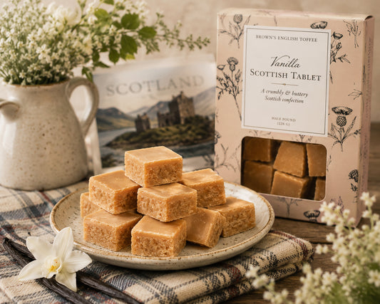 Stacked tan Scottish tablet pieces on a ceramic plate beside a branded product box, styled with flowers and a plaid cloth on a wooden surface.