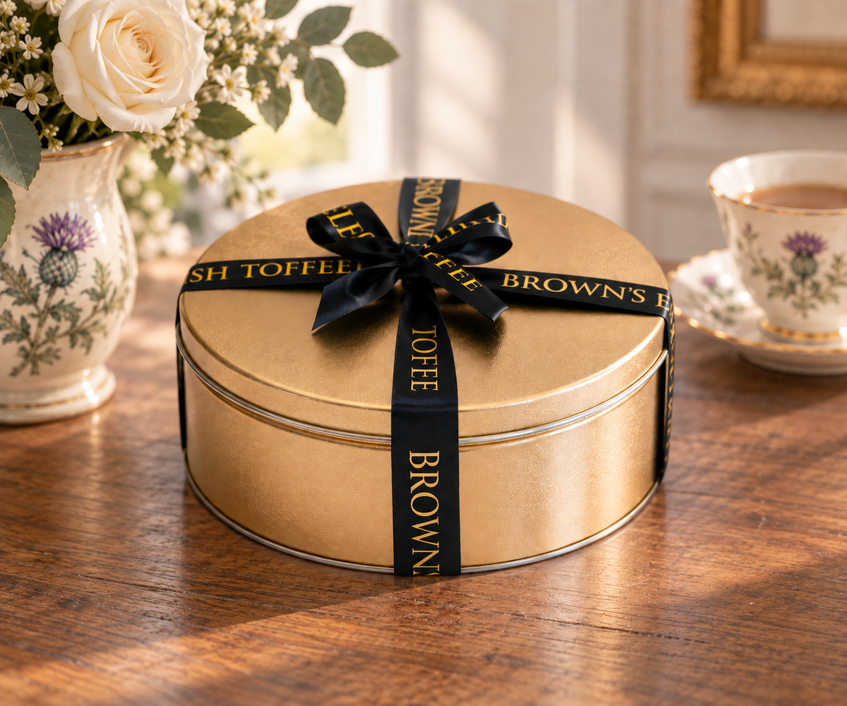 A round gold metal tin box tied with a black ribbon printed with Brown's English Toffee, set on a wooden table with a floral vase and a teacup in the background.