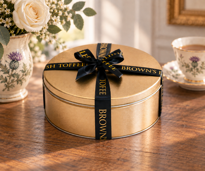 A round gold metal tin box tied with a black ribbon printed with Brown's English Toffee, set on a wooden table with a floral vase and a teacup in the background.