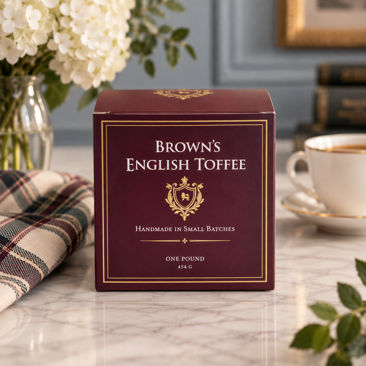 A box of Brown's English Toffee sits on a marble countertop in a styled kitchen setting, with a white teacup, flowers, and a plaid cloth nearby.