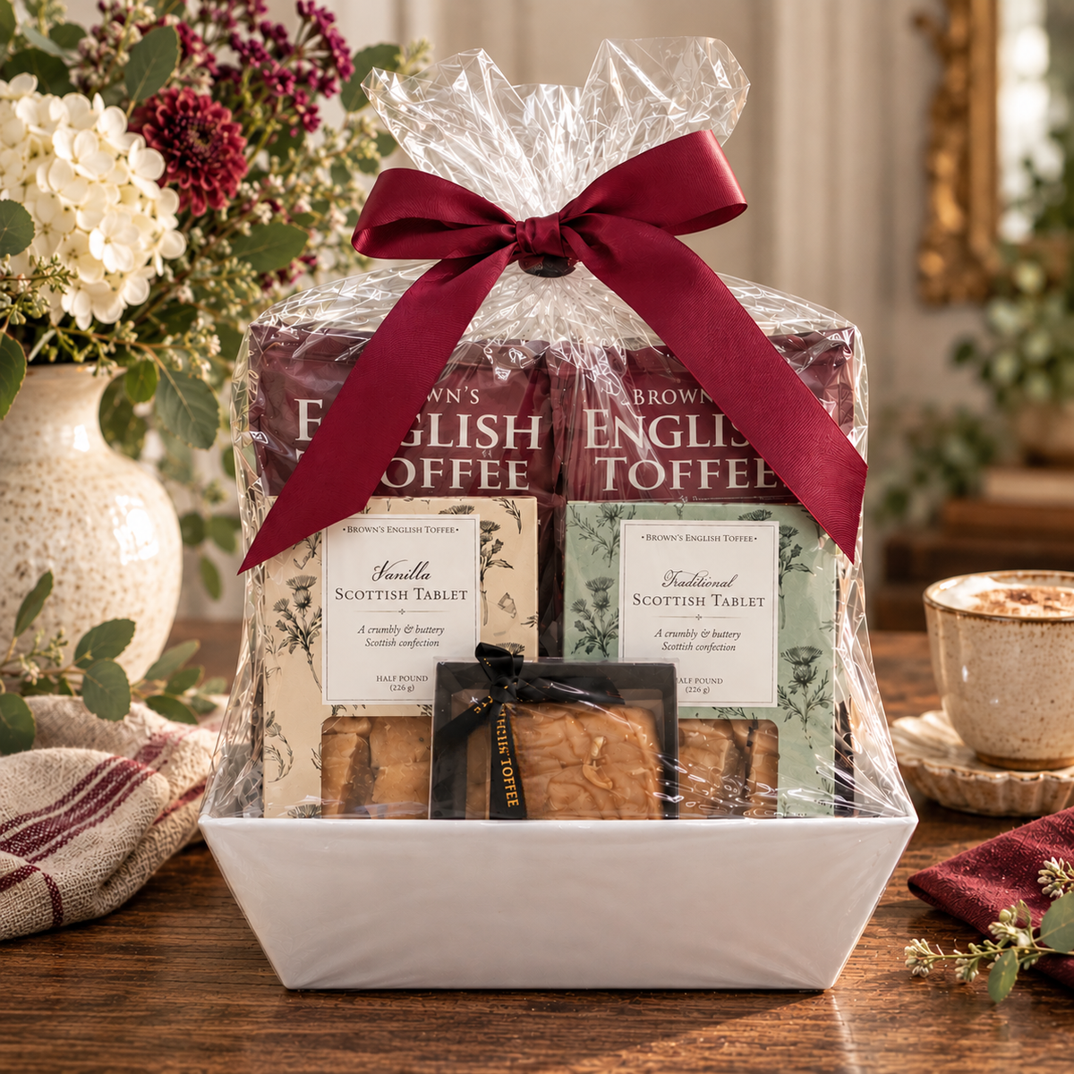 A gift basket of English Toffee, Honey Fudge and Scottish Tablet by Brown's English Toffee, wrapped in clear cellophane with a large dark red ribbon, displayed on a wooden table with a coffee cup and flowers in the background.